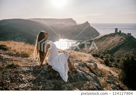A woman in a white dress is sitting on a rock overlooking a body of water. She is enjoying the view and taking in the scenery. A woman in a white dress is sitting on a rock overlooking a body of water. She is enjoying the view and taking in the scenery. 124127904
