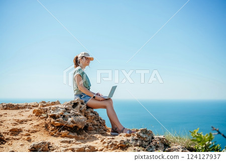 A woman is sitting on a rock overlooking the ocean with a laptop in front of her 124127937