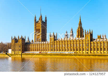 The Palace of Westminster stands majestically by the River Thames, showcasing its stunning Gothic architecture under the bright blue sky. Visitors admire its grandeur from the waterfront. 124127957