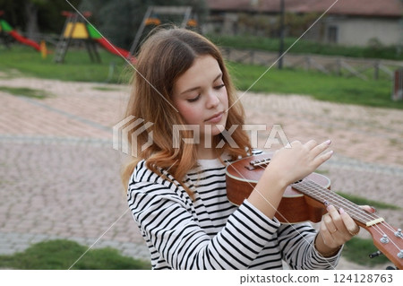 Smiling Girl Playing The Musical Instrument Ukulele In A Park Outdoor Like Violin Smiling Girl Playing The Musical Instrument Ukulele In A Park Outdoor Like Violin 124128763