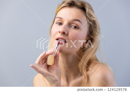 Captivating Beauty: A Glamorous Woman Applying Lip Gloss with Confidence Against a Soft Gray Background Captivating Beauty: A Glamorous Woman Applying Lip Gloss with Confidence Against a Soft Gray Background 124129576