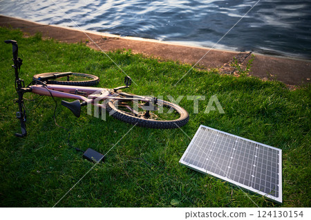 Pink electric mountain bike lies on grass near lakeside, connected to solar panel for charging. Serene scene features bike and solar panel with calm water in background. 124130154