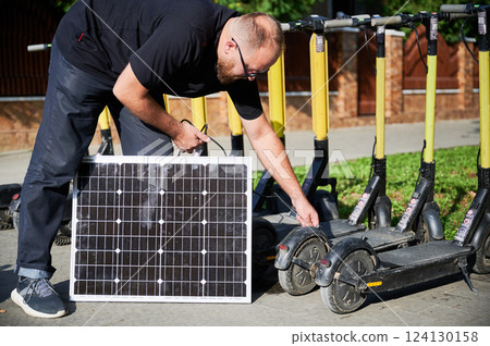 Man connects solar panel to electric scooter for charge, highlighting practical application of sustainable renewable energy. Concept of integration of eco-friendly technology in urban transportation. 124130158