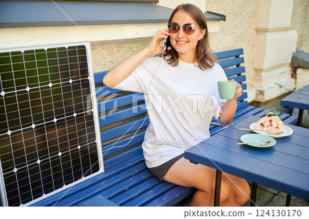 Smiling girl drinking coffee while her phone charging from solar panel. Happy woman sitting next to portable solar battery charger. Integration of sustainable renewable energy into everyday life. Smiling girl drinking coffee while her phone charging from solar panel. Happy woman sitting next to portable solar battery charger. Integration of sustainable renewable energy into everyday life. 124130170