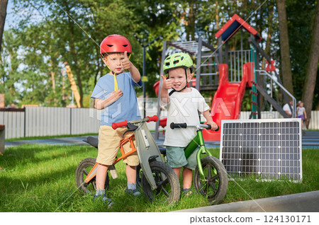 Two boys wearing helmets give thumbs-up while standing with balance bikes next to solar panel on grass. Behind them, playground with red slides and climbing structures surrounded by trees and fence. Two boys wearing helmets give thumbs-up while standing with balance bikes next to solar panel on grass. Behind them, playground with red slides and climbing structures surrounded by trees and fence. 124130171