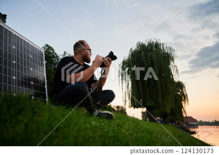 Photographer sits on grass at sunset, capturing serene landscape with camera. Solar panel set up beside, emphasizing use of renewable energy to power equipment. Sustainable practices in photography. 124130173