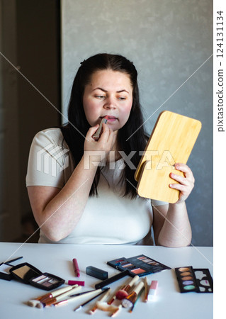 Vertical image of Millennial Caucasian woman doing self make up applying lipstick to lips while looking in mirror. Getting ready. Beauty concept. 124131134