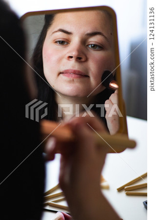 Vertical image of Millennial Caucasian woman doing self make up applying powder with make up brush looking in mirror. Reflection in mirror. Getting ready. Beauty concept. Vertical image of Millennial Caucasian woman doing self make up applying powder with make up brush looking in mirror. Reflection in mirror. Getting ready. Beauty concept. 124131136