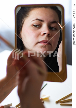 Vertical image of Millennial Caucasian woman doing self make up painting eyebrows looking in mirror. Reflection in mirror. Getting ready. Beauty concept. Vertical image of Millennial Caucasian woman doing self make up painting eyebrows looking in mirror. Reflection in mirror. Getting ready. Beauty concept. 124131137