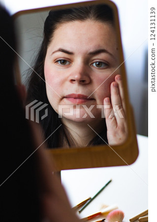Vertical image of Millennial Caucasian woman doing self make up applying face cream while looking in mirror. Reflection in mirror. Getting ready. Beauty concept. Vertical image of Millennial Caucasian woman doing self make up applying face cream while looking in mirror. Reflection in mirror. Getting ready. Beauty concept. 124131139