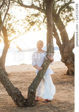 Blonde teenage girl in white shirt standing near tree on the background of sea and sunset Blonde teenage girl in white shirt standing near tree on the background of sea and sunset 124131498