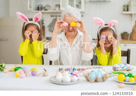 Happy Easter. Two little girls sisters twins grandmother painting eggs enjoying time together. Happy family grandma granddaughters child kids preparing for Easter. Spring Christian festival tradition 124133010