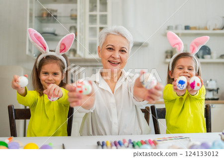 Happy Easter. Two little girls sisters twins grandmother painting eggs enjoying time together. Happy family grandma granddaughters child kids preparing for Easter. Spring Christian festival tradition Happy Easter. Two little girls sisters twins grandmother painting eggs enjoying time together. Happy family grandma granddaughters child kids preparing for Easter. Spring Christian festival tradition 124133012