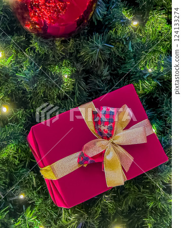 Beautifully wrapped Christmas gift with a golden ribbon placed under a decorated tree with glowing festive lights 124133274
