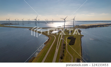 A scenic aerial view of a coastal wind farm with multiple wind turbines standing over the water. Curved roads lead through the landscape, with cars visible. The sun reflects off the water. 124133453