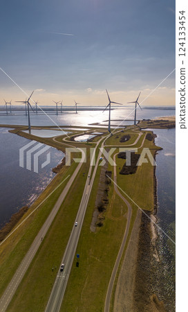 A scenic aerial view of a coastal wind farm with multiple wind turbines standing over the water. Curved roads lead through the landscape, with cars visible. The sun reflects off the water. A scenic aerial view of a coastal wind farm with multiple wind turbines standing over the water. Curved roads lead through the landscape, with cars visible. The sun reflects off the water. 124133454