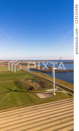 A scenic aerial view of a coastal wind farm with multiple wind turbines standing over the water. Curved roads lead through the landscape, with cars visible. The sun reflects off the water. A scenic aerial view of a coastal wind farm with multiple wind turbines standing over the water. Curved roads lead through the landscape, with cars visible. The sun reflects off the water. 124133456