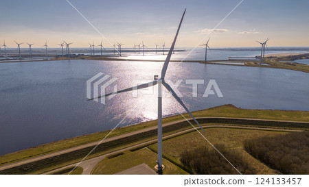 A large wind turbine stands near a coastal waterway, reflecting sunlight. More turbines line the horizon, generating renewable energy. Curved roads and green fields complete this scenic. 124133457