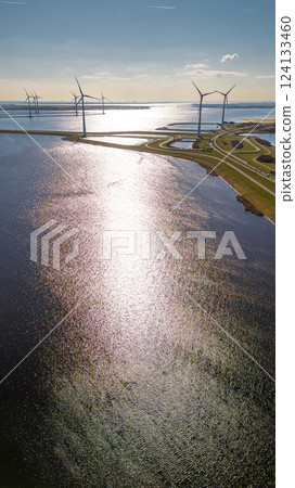 A scenic aerial view of a coastal wind farm with multiple wind turbines standing over the water. Curved roads lead through the landscape, with cars visible. The sun reflects off the water. 124133460