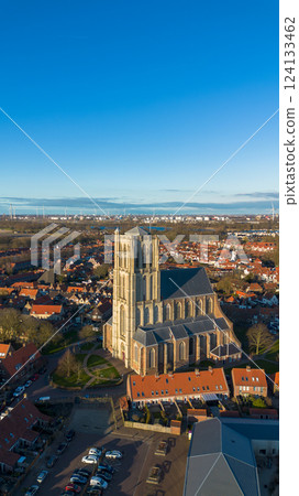 Aerial view of a historic town with a tall Gothic church in the center. Red-roofed houses, narrow streets, and a distant industrial skyline create a mix of old and modern architecture. 124133462