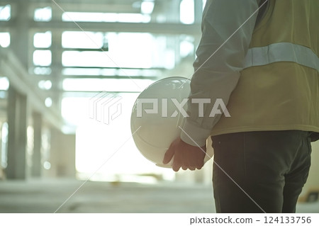 Construction engineer holding hardhat inspecting building site during bright day 124133756
