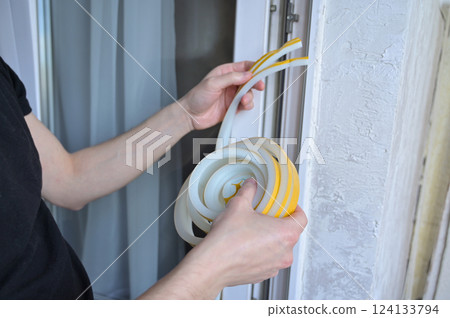 A man insulates windows and balcony doors with foam adhesive tape. Budget renovation in the apartment. Preparation for the cold snap. Heating problem in apartment buildings 124133794