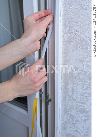 A man's hands insulates windows and balcony doors with foam adhesive tape. Close up. Budget renovation in the apartment. Preparation for the cold snap. Heating problem in apartment buildings 124133797