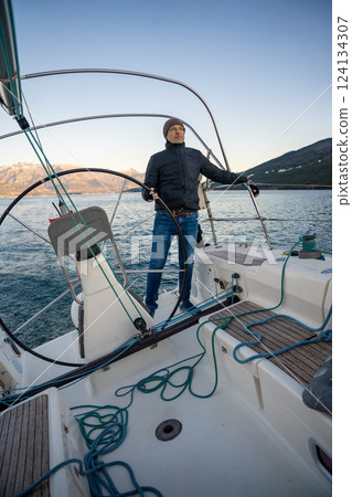 Young man captain standing at the helm and controls a sailboat during a journey by sea in winter time in Montenegro Young man captain standing at the helm and controls a sailboat during a journey by sea in winter time in Montenegro 124134307