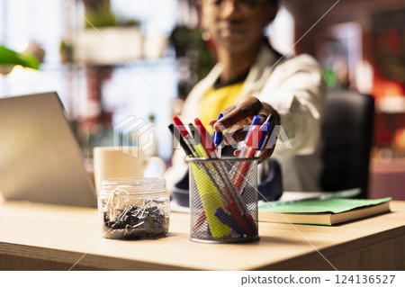 Woman reaches for pen from her desk organizer, preparing to annotate document after brainstorming ideas. Student in home office requiring writing tool from stationary items holder, close up 124136527