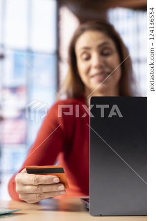 Adult holds her card to check balance and oversee budget planning, uses laptop at home office to track her expenses responsibly and confirm payments. Mature woman manages her finances. 124136554