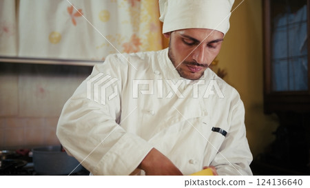 Italian Man Chef In Uniform Peels Potatoes On Wooden Cutting Board At His Home Italian Man Chef In Uniform Peels Potatoes On Wooden Cutting Board At His Home 124136640