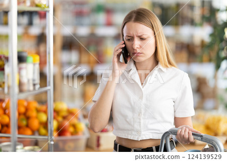 Upset young woman choosing groceries while talking on phone Upset young woman choosing groceries while talking on phone 124139529