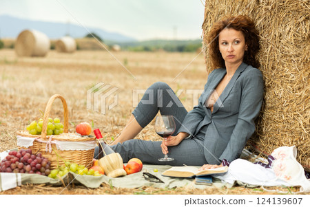 Woman in unbuttoned jacket with bare chest sits near hay bale on picnic 124139607