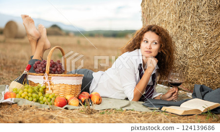 Relaxed businesswoman lying near haystack with wine and book 124139686