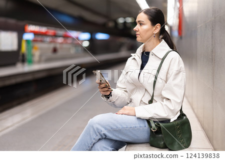 Girl with phone is sitting on metro platform station, scrolling though internet 124139838