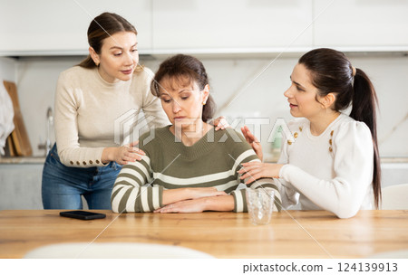 Sad woman sits silently in kitchen, adult girls sympathize and comfort her Sad woman sits silently in kitchen, adult girls sympathize and comfort her 124139913