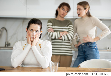 Upset middle-aged woman sitting at the kitchen table with her back to women quarreling to her Upset middle-aged woman sitting at the kitchen table with her back to women quarreling to her 124140072