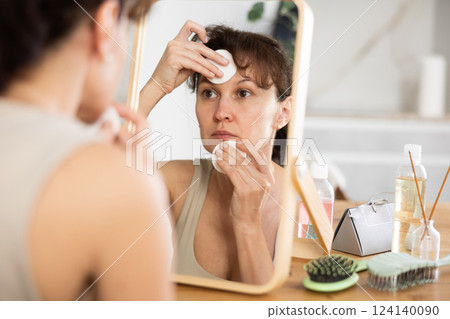 Woman looking in mirror removing cosmetics form face with cotton rounds 124140090