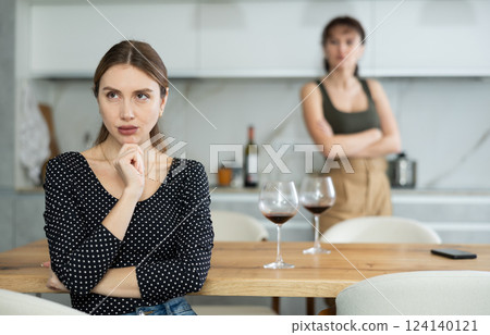 Upset middle-aged woman sitting at the kitchen table with her back to another lady quarreling to her 124140121