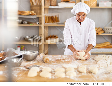 Aged female baker kneading dough on floured table in bakery 124140139