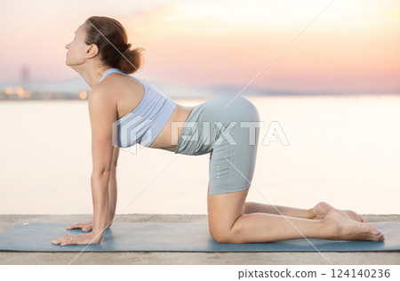 Skilled young female trainer performing cat-cow pose of yoga on seashore at sunrise Skilled young female trainer performing cat-cow pose of yoga on seashore at sunrise 124140236