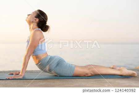 Sportive woman performing cobra pose of yoga during training on beach at sunrise Sportive woman performing cobra pose of yoga during training on beach at sunrise 124140392