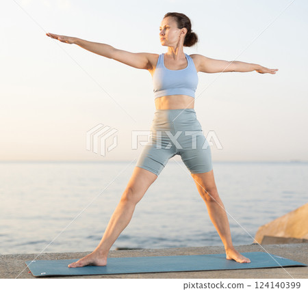 Near sea during sunset, girl performs variations of virabhadrasana 124140399