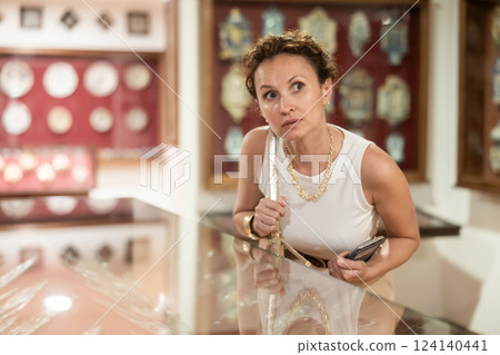 Woman with paper and phone in hands walks around hall of museum, examines showpiece at exhibition 124140441