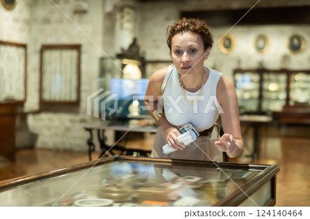 Woman with paper and phone in hands walks around hall of museum, examines showpiece at exhibition 124140464