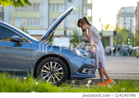 Helpless woman standing near her car with open hood takling on mobile phone calling road service for help. Young female driver having trouble with vehicle. 124140766