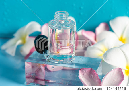 A glistening serum in a dropper bottle sits on a clear block with fresh flowers nearby. 124143216