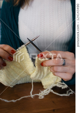 Women hands holding knitting needles and yellow cotton knitted work. Knitting woman with bobbins of yarn and yellow knitwear. Needlework in process. Knitting hobby. Knitter work. Women hands holding knitting needles and yellow cotton knitted work. Knitting woman with bobbins of yarn and yellow knitwear. Needlework in process. Knitting hobby. Knitter work. 124143490