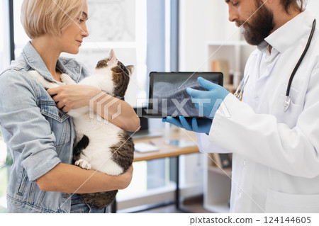 Veterinarian in clinic explains x-ray results to young adult female holding pet cat. Image portrays veterinary care, animal health, compassion, and medical attention focused on a domestic cat. Veterinarian in clinic explains x-ray results to young adult female holding pet cat. Image portrays veterinary care, animal health, compassion, and medical attention focused on a domestic cat. 124144605