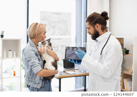 Veterinarian explaining X-ray image to young woman holding her pet cat indoors office. Image suitable for health care, veterinarian services, and animal welfare. 124144607